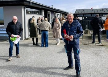 Campingfatter Tor Ove Sørlie med Dannebrog under artmen og Jens-Arne Nielsen i hælene klar til standerhejsning hos Hirtshals Camping. I baggrunden ses formand Sarah Routledge og med ryggen til turistchef Laila Zielke fra Hirtshals Turisme. Foto: Hirtshals Camping.
