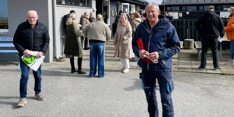 Campingfatter Tor Ove Sørlie med Dannebrog under artmen og Jens-Arne Nielsen i hælene klar til standerhejsning hos Hirtshals Camping. I baggrunden ses formand Sarah Routledge og med ryggen til turistchef Laila Zielke fra Hirtshals Turisme. Foto: Hirtshals Camping.